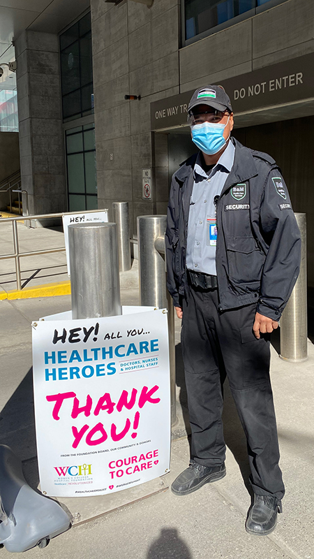 A security guard with a mask stands out of the parking garage of the hospital, beside a sign that says "Hey Health Care Heroes, Thank You!"