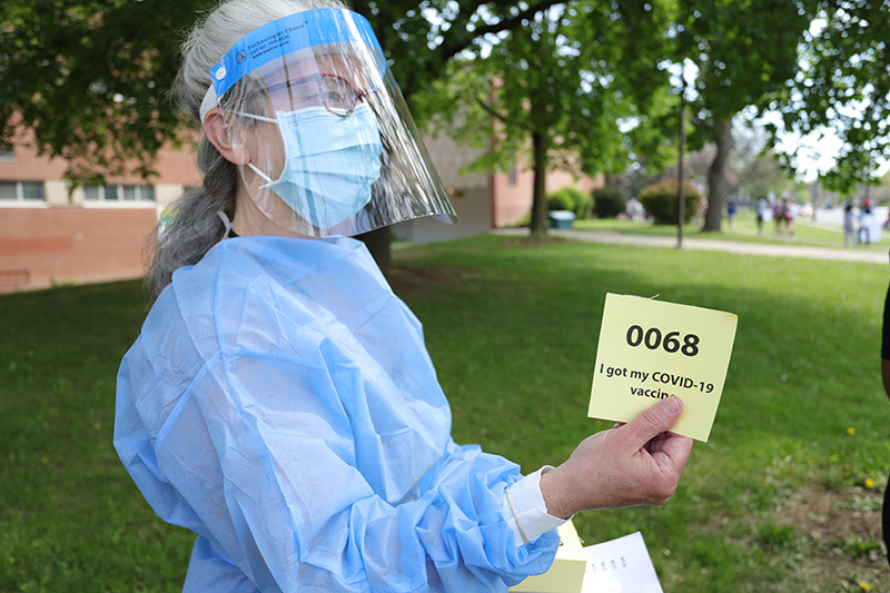 A woman in full PPE gear holds a ticket that reads "I got my COVID-19 Vaccine"